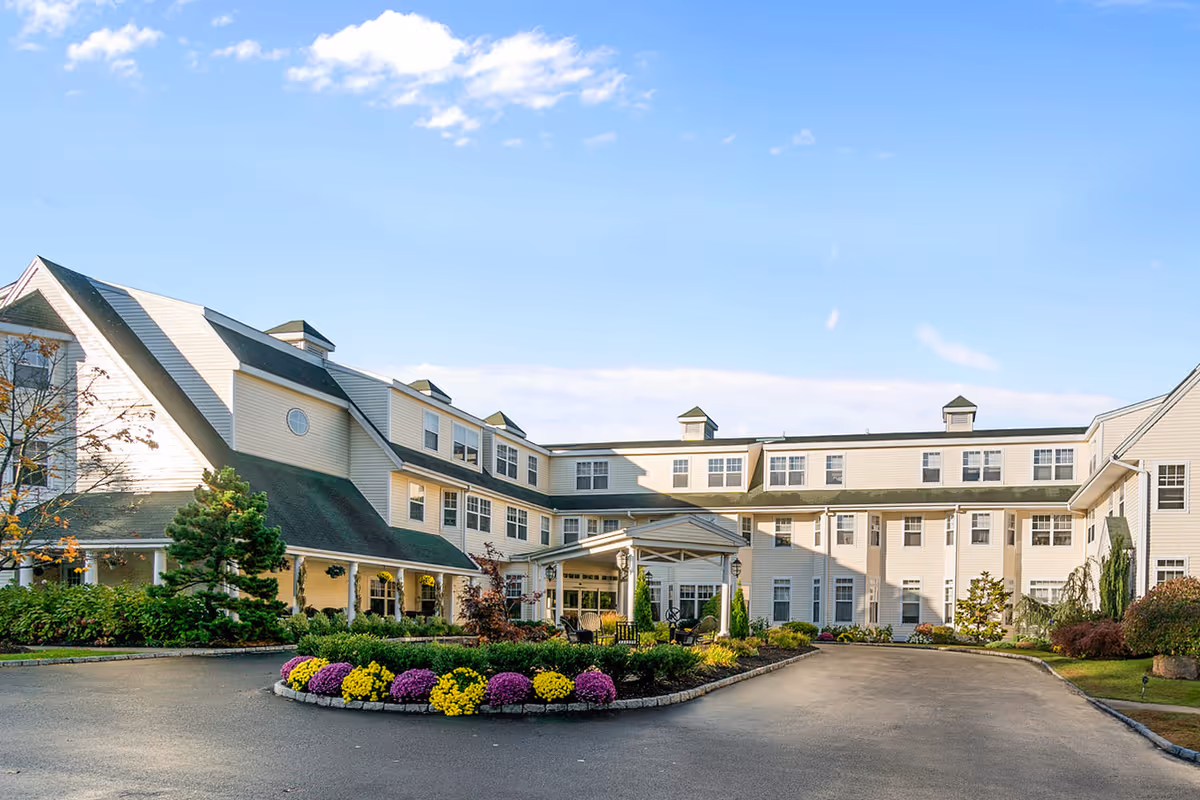 Front exterior of a large three-story senior living facility with a circular driveway, landscaped flowerbed and covered main entrance under a blue sky.