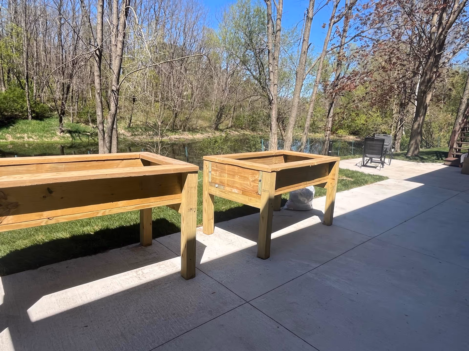 Outdoor patio area with two wooden raised garden beds on a concrete surface. In the background, there are trees with sparse leaves and a small body of water. A black chair is visible near the edge of the patio under a clear blue sky.