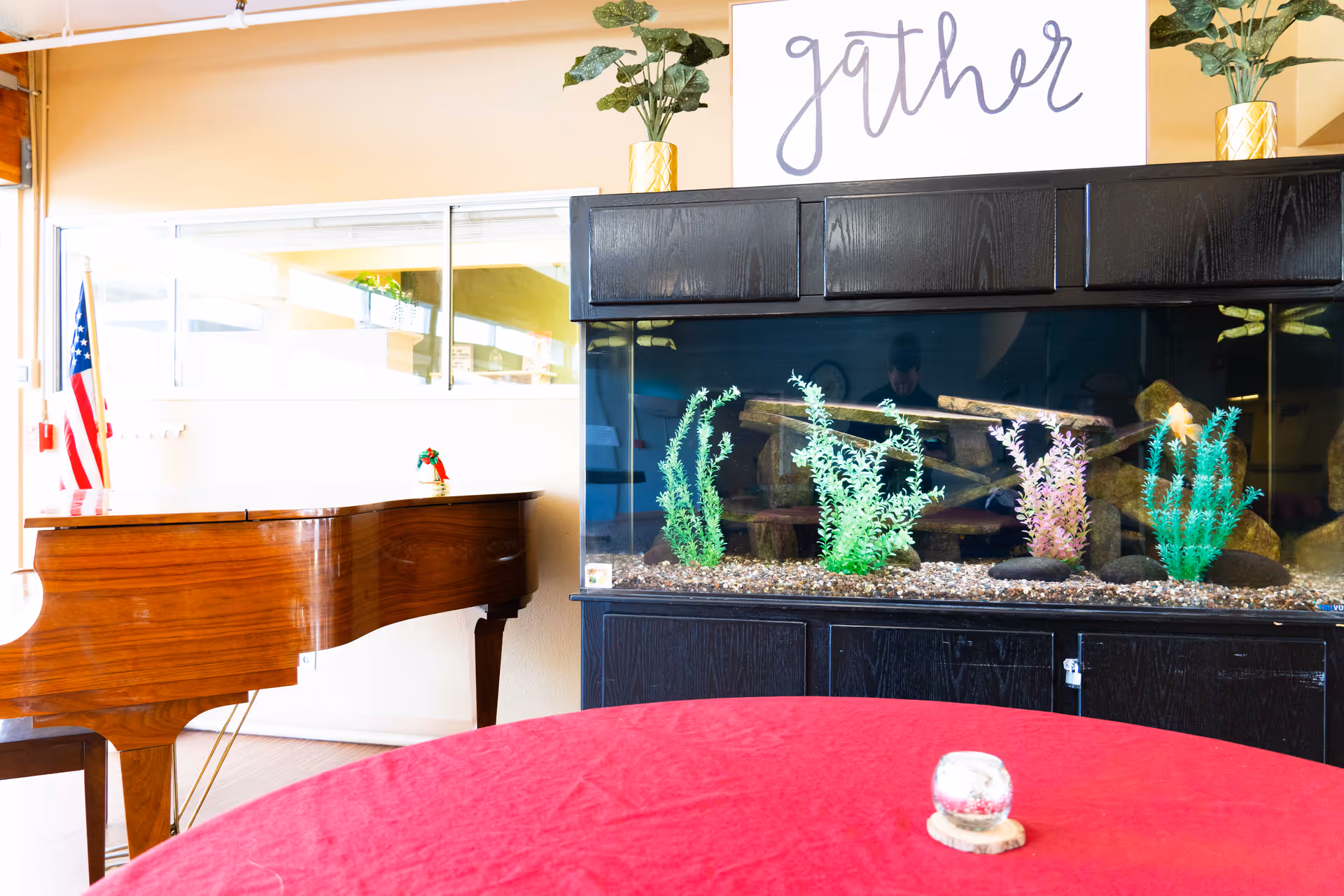 Interior room with a large fish tank decorated with artificial plants, a wooden grand piano, a round table covered with a red tablecloth, and a sign above the fish tank that says 'gather'. There are two potted plants on top of the fish tank cabinet and an American flag in the background.