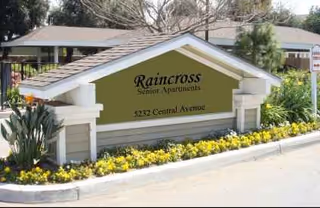A Raincross Senior Apartments sign sits at a landscaped entrance with yellow flowers.
