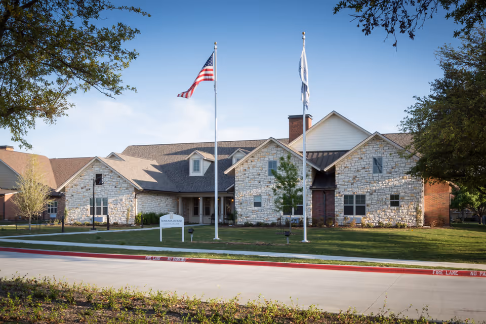 Exterior view of Sonoma House Assisted Living Alzheimer’s Care building with stone and brick facade, two flagpoles with American and white flags, green lawn, and trees under a clear blue sky.