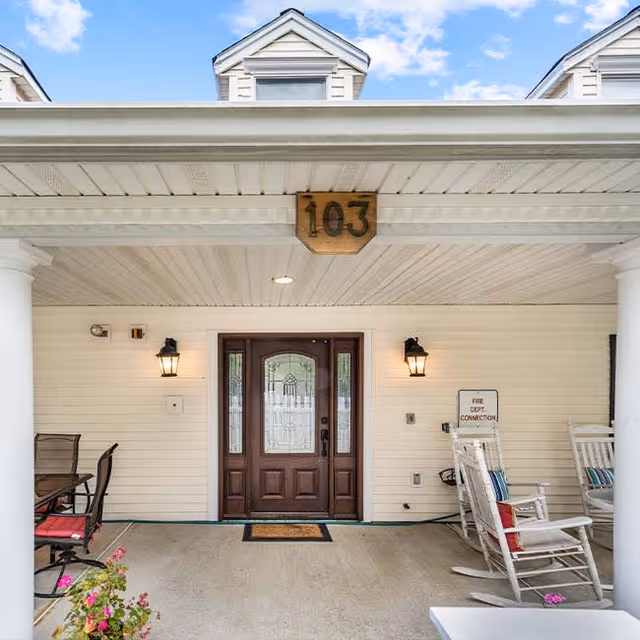 Entrance to a building with a wooden door featuring decorative glass panels, flanked by two wall-mounted lantern-style lights. The porch area has white columns, a ceiling with recessed lighting, and a wooden sign above the door displaying the number 103. There are rocking chairs with cushions on the right side and a table with chairs on the left side, along with some potted flowers near the edge of the porch.