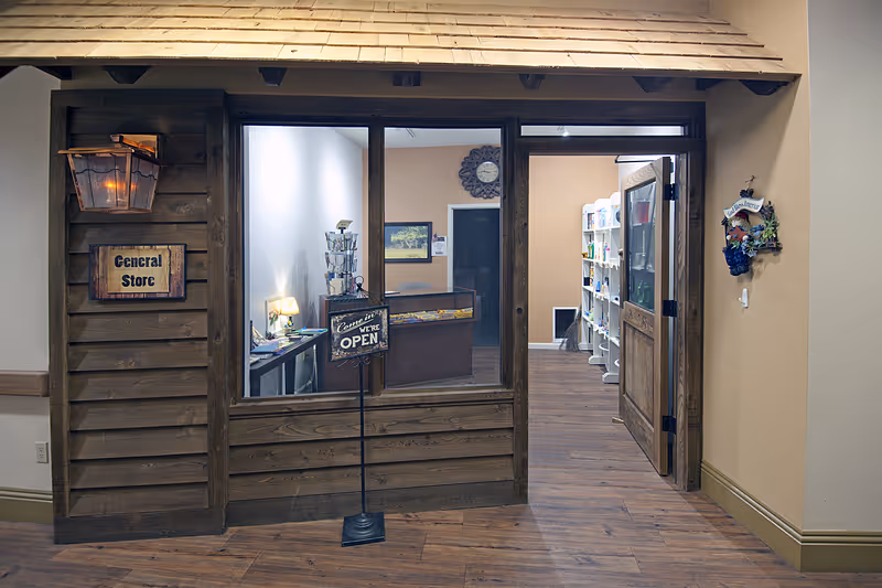 Interior view of a small mock 'General Store' storefront in a senior living facility hallway, showing a wooden facade, an 'Open' sign, and shelves visible through the window.