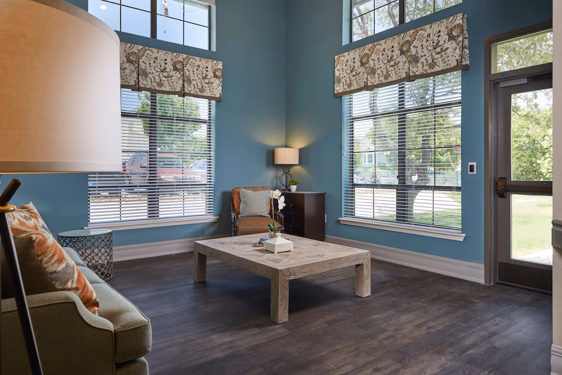 A cozy living room with teal walls, large windows with floral valances, a wooden coffee table with a small plant and books, a green sofa with patterned pillows, an armchair with a pillow, a side table with a lamp, and a glass door leading outside.