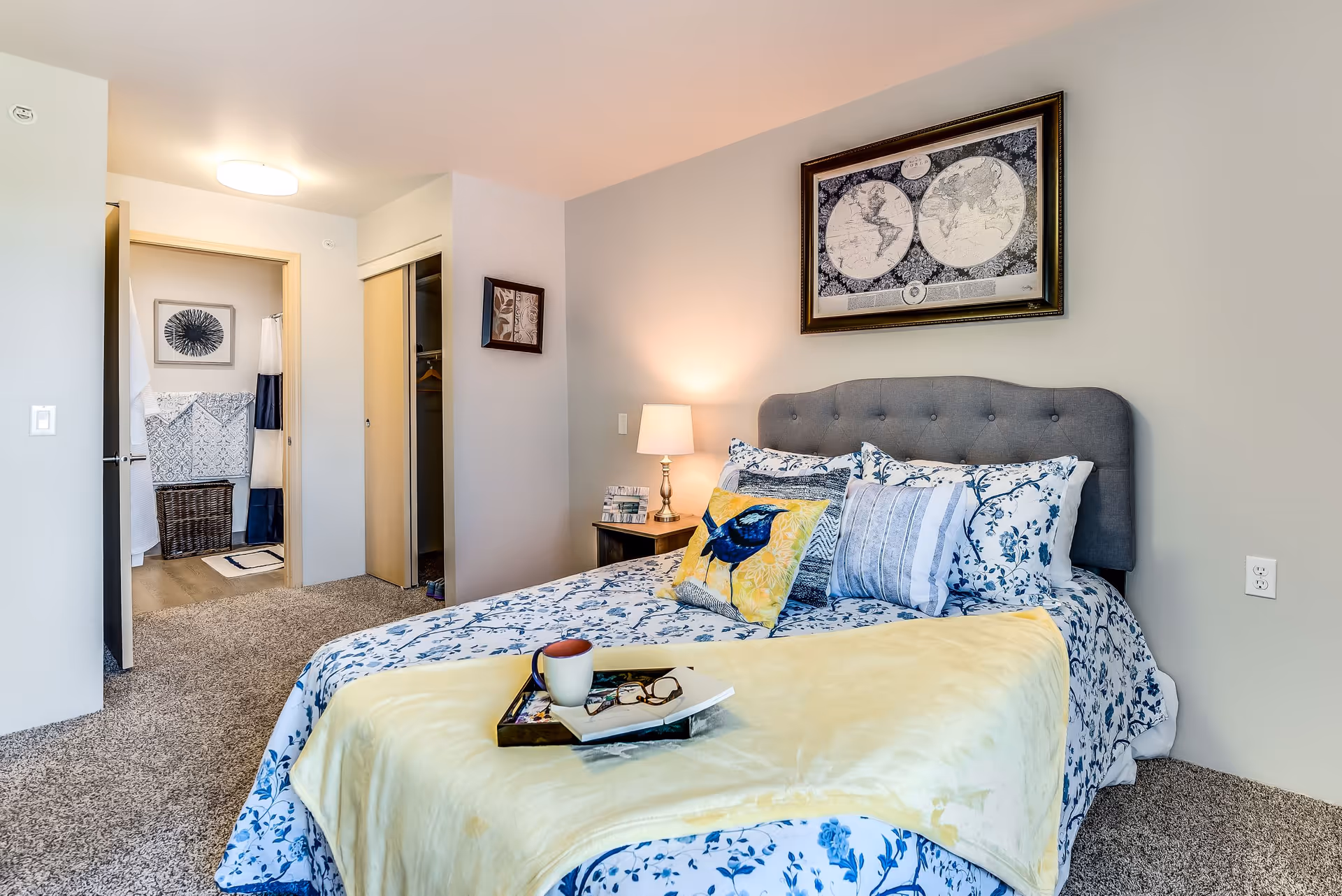 A cozy bedroom with a bed featuring a gray tufted headboard, floral bedding, and multiple pillows including one with a bird design. A yellow throw blanket is draped over the bed with a tray holding a cup, glasses, and a book. A nightstand with a lamp and books is beside the bed. The room has beige walls, carpeted floor, and a framed world map hanging above the bed. An open doorway leads to a bathroom with a shower curtain and a laundry basket visible.