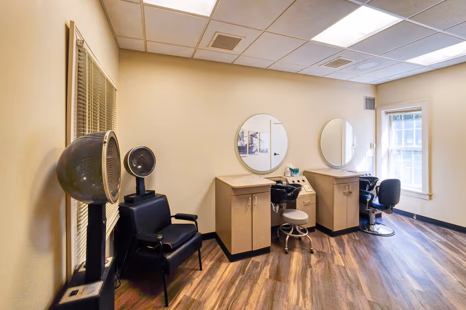 Interior of a hair salon area in a senior living facility with two hair washing stations, two round mirrors mounted on the wall, salon chairs, and hair dryers. The room has wood-patterned flooring, beige walls, and a window letting in natural light.