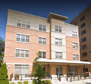 Exterior view of McAuley Apartments, a multi-story residential building with a combination of light brick and white paneling. The entrance features a covered porch with railings and a small landscaped area with plants and a tree in front.