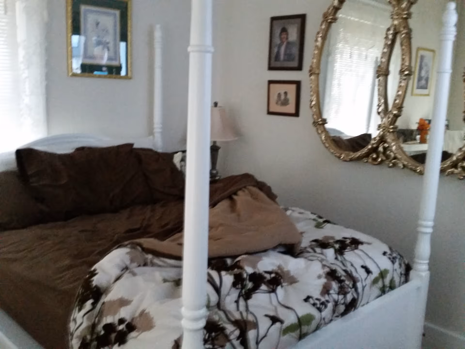 A bedroom with a white four-poster bed featuring a floral patterned comforter and brown pillows. The room has a lamp on a bedside table, framed pictures on the walls, and an ornate gold-framed mirror reflecting part of the room.