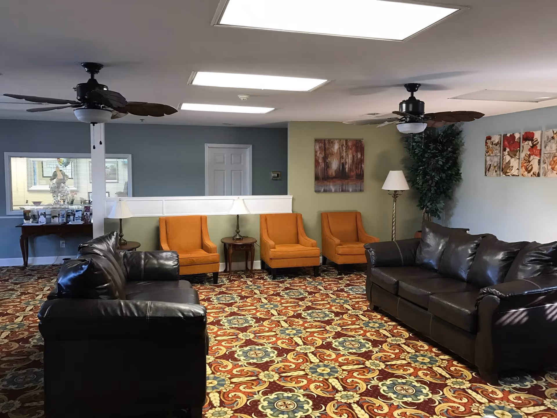 A cozy living room area in Willow Creek Assisted Living featuring two dark brown leather sofas facing each other, three orange armchairs lined up against a green accent wall, two ceiling fans with leaf-shaped blades, a patterned carpet with red, yellow, and blue designs, table lamps on small wooden tables, a floor lamp, and wall art including a painting of trees and floral prints.