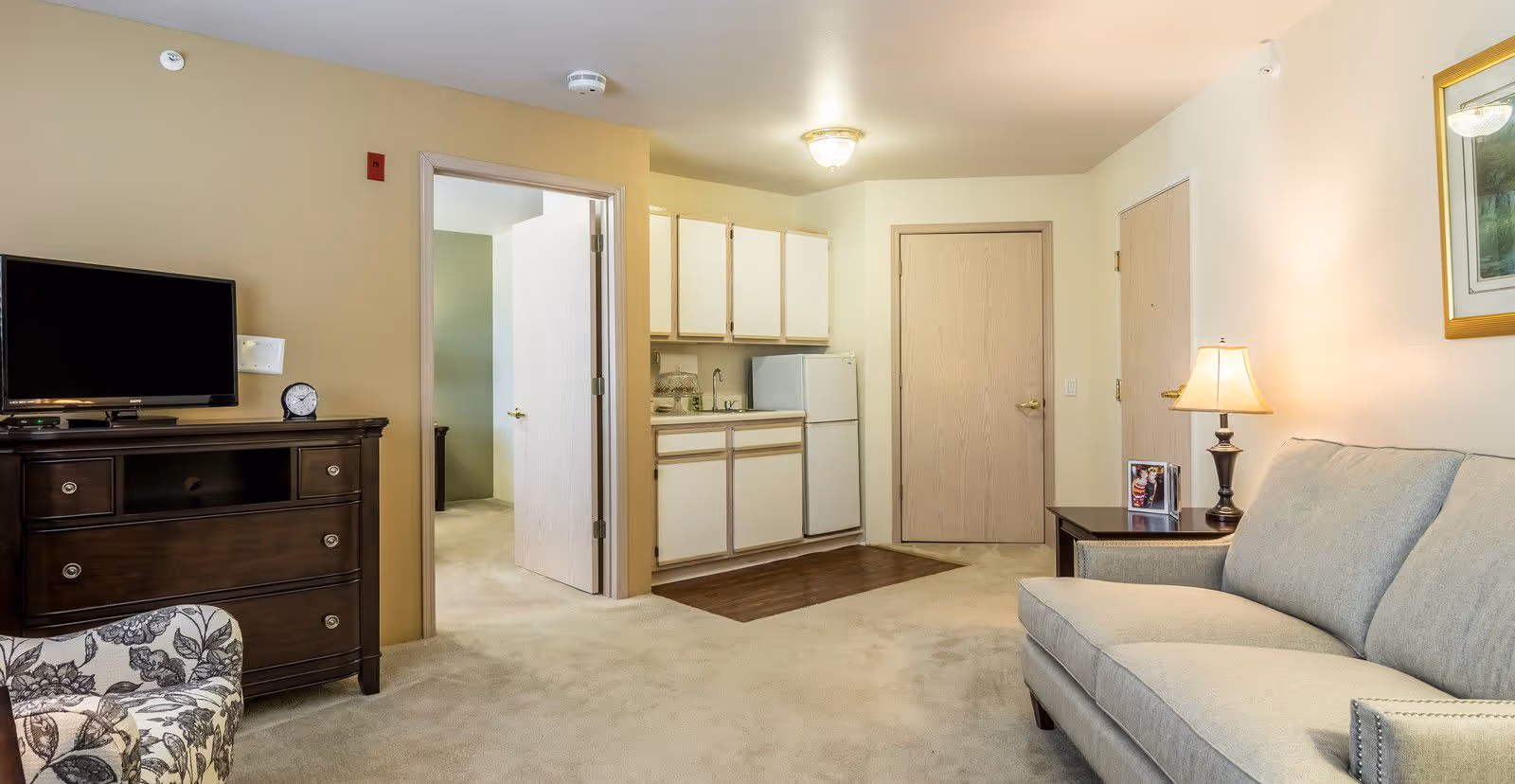A cozy living room area with a light gray sofa, a side table with a lamp and a framed photo, a floral patterned armchair, and a dark wooden TV stand with a flat-screen TV and a clock. In the background, there is a small kitchenette with white cabinets, a sink, and a refrigerator. Two doors are visible, one open leading to another room and one closed.