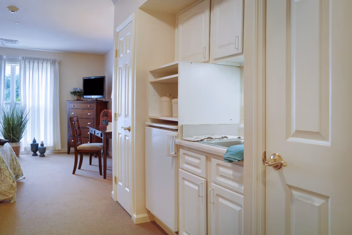 Interior view of a senior living facility room at Sunrise of Findlay showing a small kitchenette with white cabinets and a mini fridge on the right, and a living area with a wooden chair, a small table, a chest of drawers with a TV on top, and a window with white curtains letting in natural light on the left.