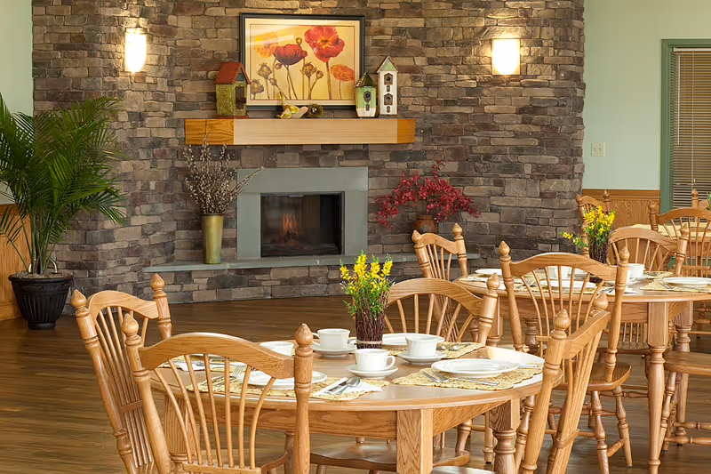 Dining area with wooden tables and chairs set with white cups, plates, and cutlery. A stone fireplace with a wooden mantle is in the background, decorated with a framed floral painting, small birdhouses, and vases with flowers. The room has wooden flooring and soft lighting from wall sconces.