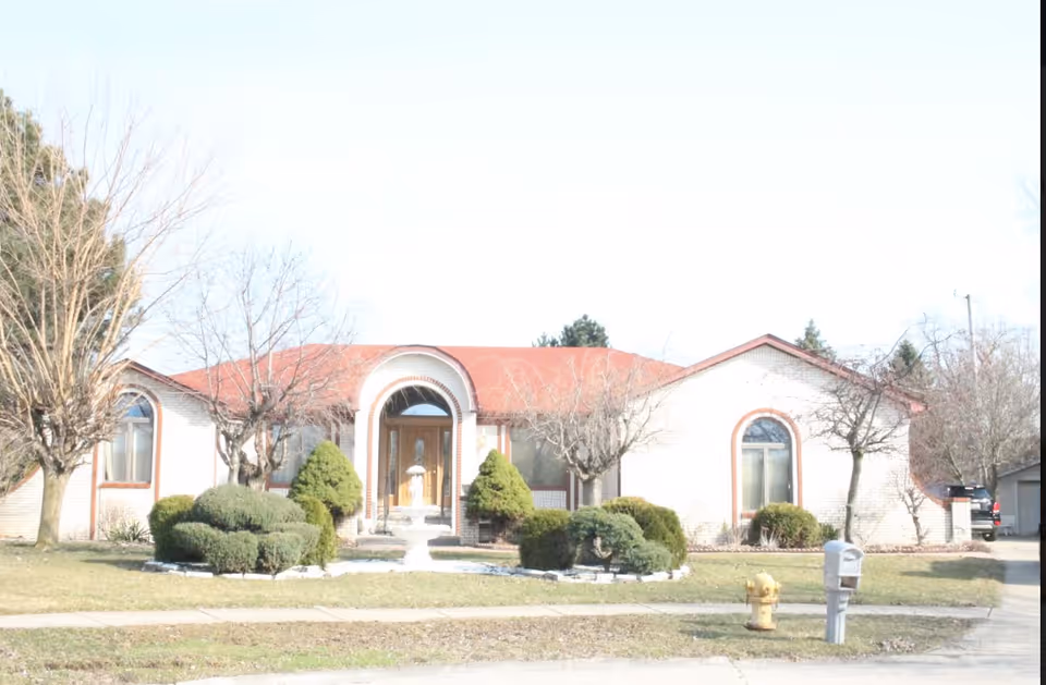 Front exterior view of a single-story building with a red roof and arched windows. The entrance features a wooden door with a decorative archway. There are trimmed bushes and a small fountain in the front yard, along with leafless trees and a mailbox near the sidewalk.