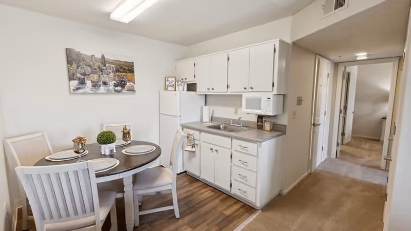 A small kitchen and dining area in a senior living facility. The kitchen has white cabinets, a countertop with a sink, a microwave, and a refrigerator. The dining area features a round wooden table set with four chairs, placemats, and decorative items. The floor is a combination of wood and carpet, and a hallway with doors is visible in the background.