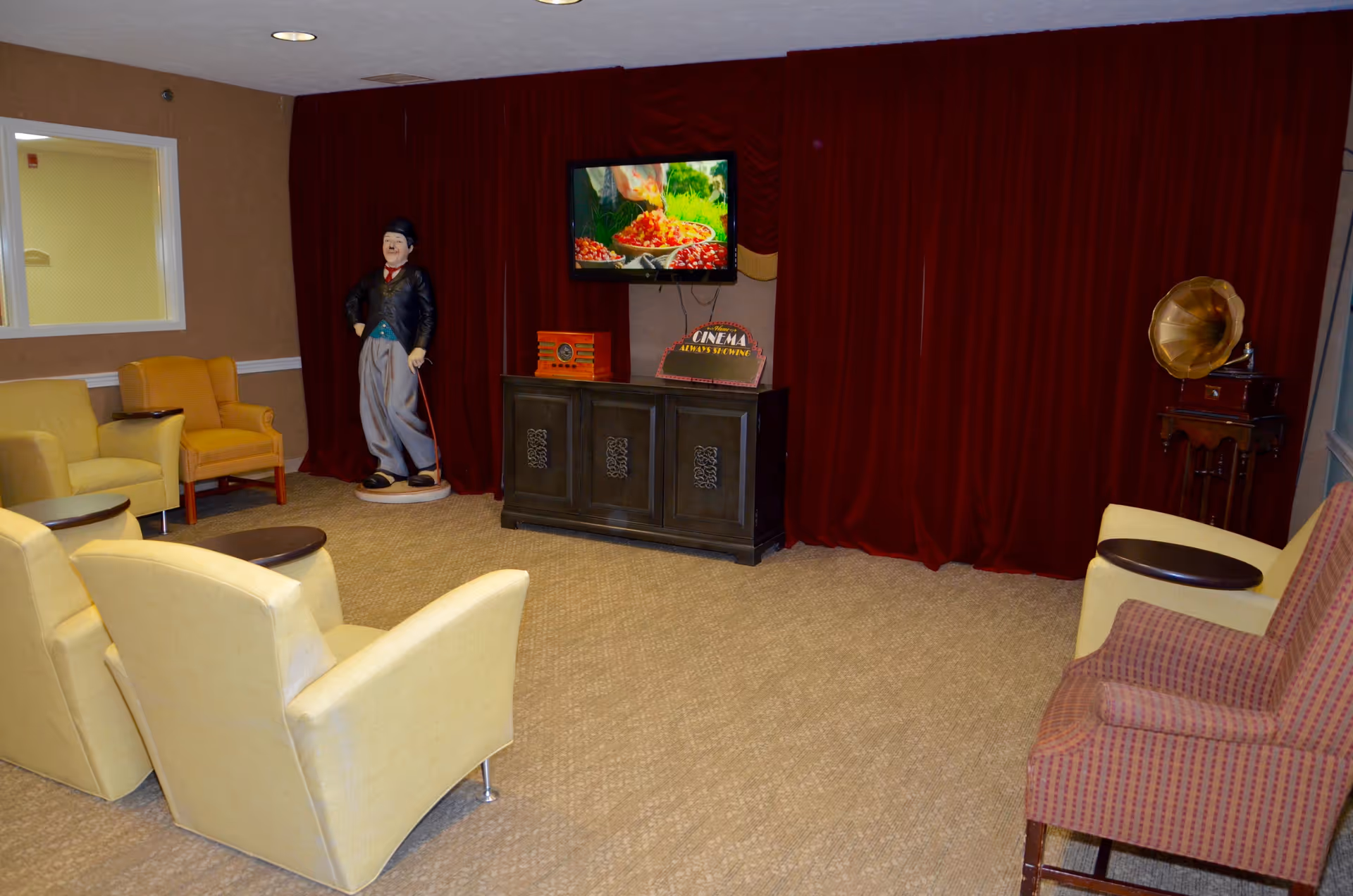 A cozy living room area with several upholstered chairs arranged facing a TV mounted on a dark wooden cabinet. The room features a beige carpet, red curtains covering the back wall, a vintage gramophone on a small wooden stand, and a life-sized statue of Charlie Chaplin standing near the TV.
