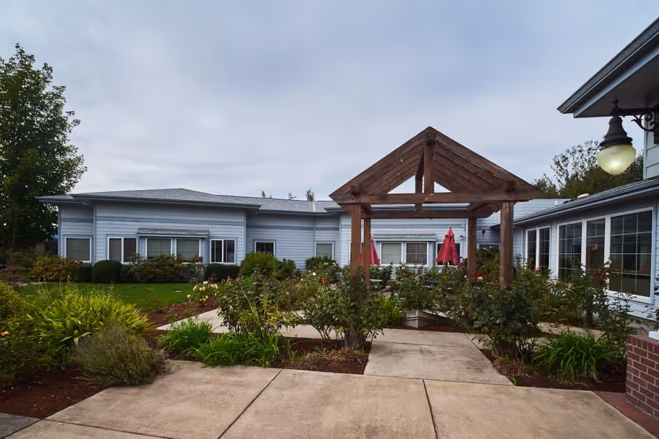 Outdoor courtyard area of a senior living facility with a wooden pergola structure in the center, surrounded by green bushes and plants. The building has light gray siding with multiple windows and red umbrellas visible in the background. A concrete pathway leads through the garden area under the pergola.