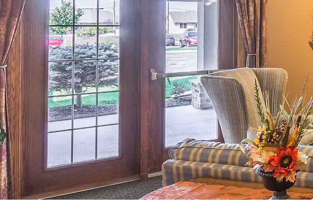 View of a cozy seating area inside a room with a striped armchair and a matching sofa near large wooden-framed glass doors that open to an outdoor patio. A floral arrangement in a vase is on a table in the foreground, and outside there is a sidewalk, green grass, bushes, and parked cars.