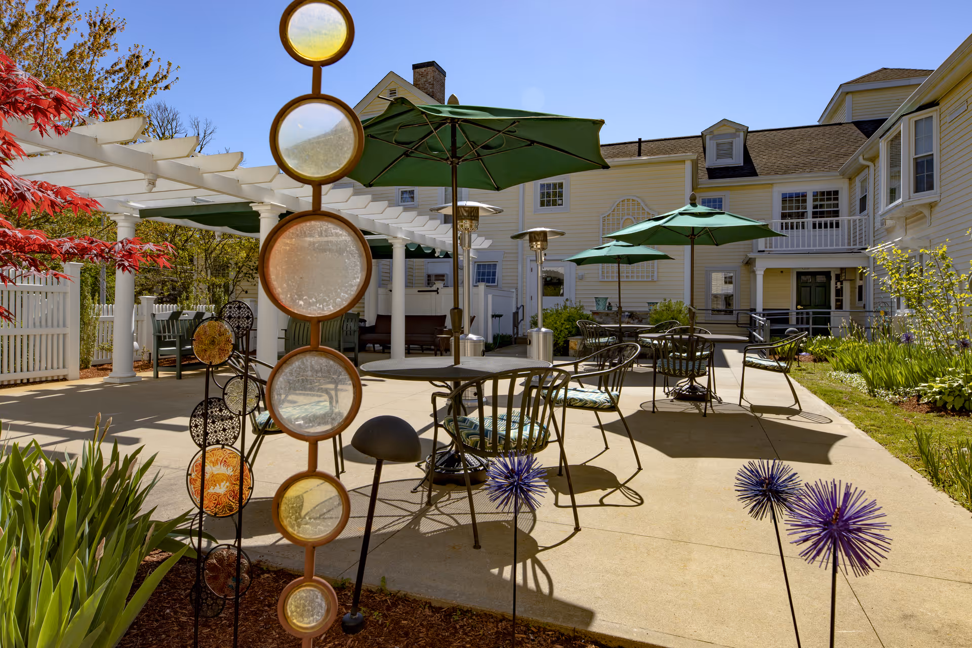 Outdoor patio area at a senior living facility with metal tables and chairs, green umbrellas providing shade, decorative garden sculptures, and a white pergola. The building in the background is light yellow with multiple windows and a balcony. There are plants and flowers around the patio under a clear blue sky.