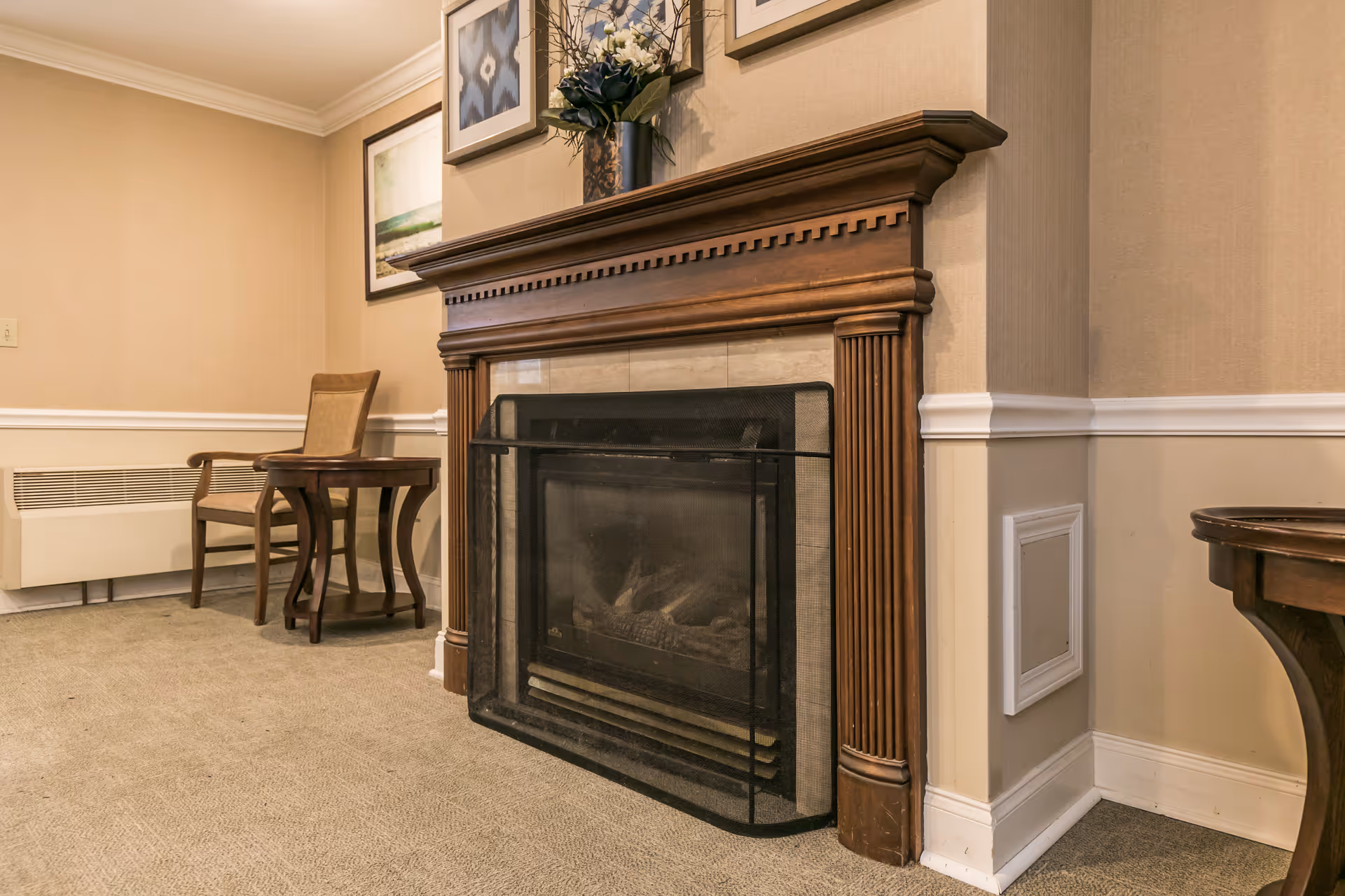 A cozy interior corner featuring a wooden fireplace with a protective screen, a small wooden side table with a chair, and framed artwork on the beige walls above the fireplace. The room has carpeted flooring and white trim along the walls.