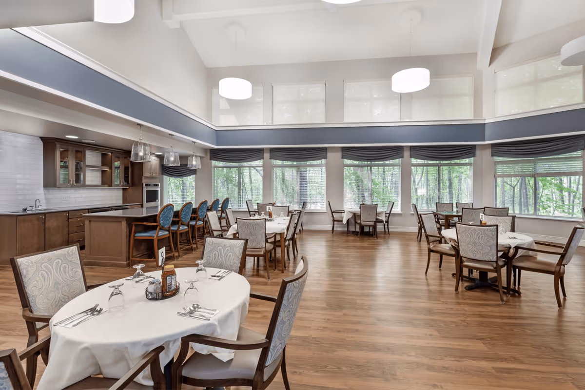 A spacious dining room in a senior living facility with multiple round tables covered with white tablecloths, each set with silverware, glasses, and condiments. The room features large windows with views of trees outside, wooden flooring, and a kitchen area with a counter and bar stools in the background. The ceiling is high with modern pendant lights.