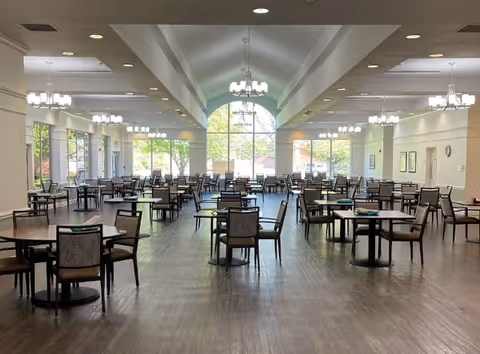 A spacious dining room with multiple round tables and chairs arranged neatly. Large windows at the far end allow natural light to fill the room. Several chandeliers hang from the ceiling, providing additional lighting. The floor is wooden, and the walls are painted white, creating a bright and welcoming atmosphere.