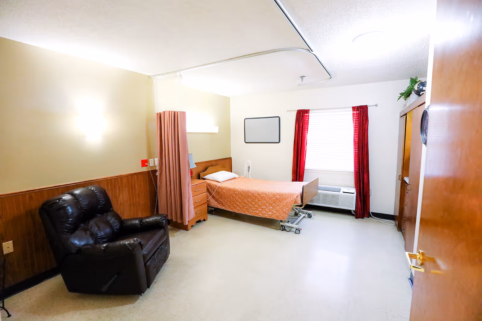 A simple nursing facility bedroom with a single hospital bed covered in an orange quilt, a black recliner chair, a wooden dresser, red curtains on a window, and a ceiling-mounted curtain track for privacy.