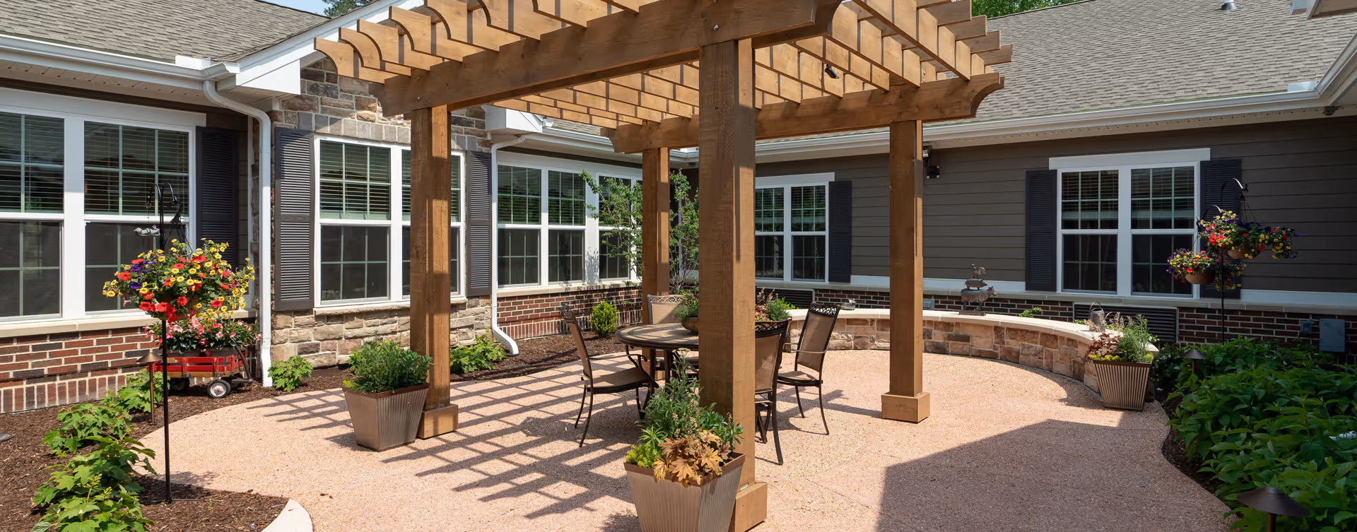 Outdoor patio area at Bickford of Chesapeake featuring a wooden pergola with a round table and four chairs underneath. The patio is surrounded by flower beds with various plants and flowers, and the building exterior has multiple windows with white frames and dark shutters.