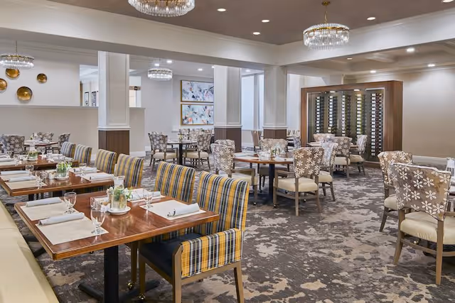 A spacious and well-lit dining room with multiple wooden tables set with placemats, napkins, glasses, and small flower arrangements. The chairs have different upholstery patterns, including striped and floral designs. The room features modern chandeliers, decorative wall art, and a wine rack against the far wall. The carpet has a floral pattern, and the walls are painted in neutral tones.