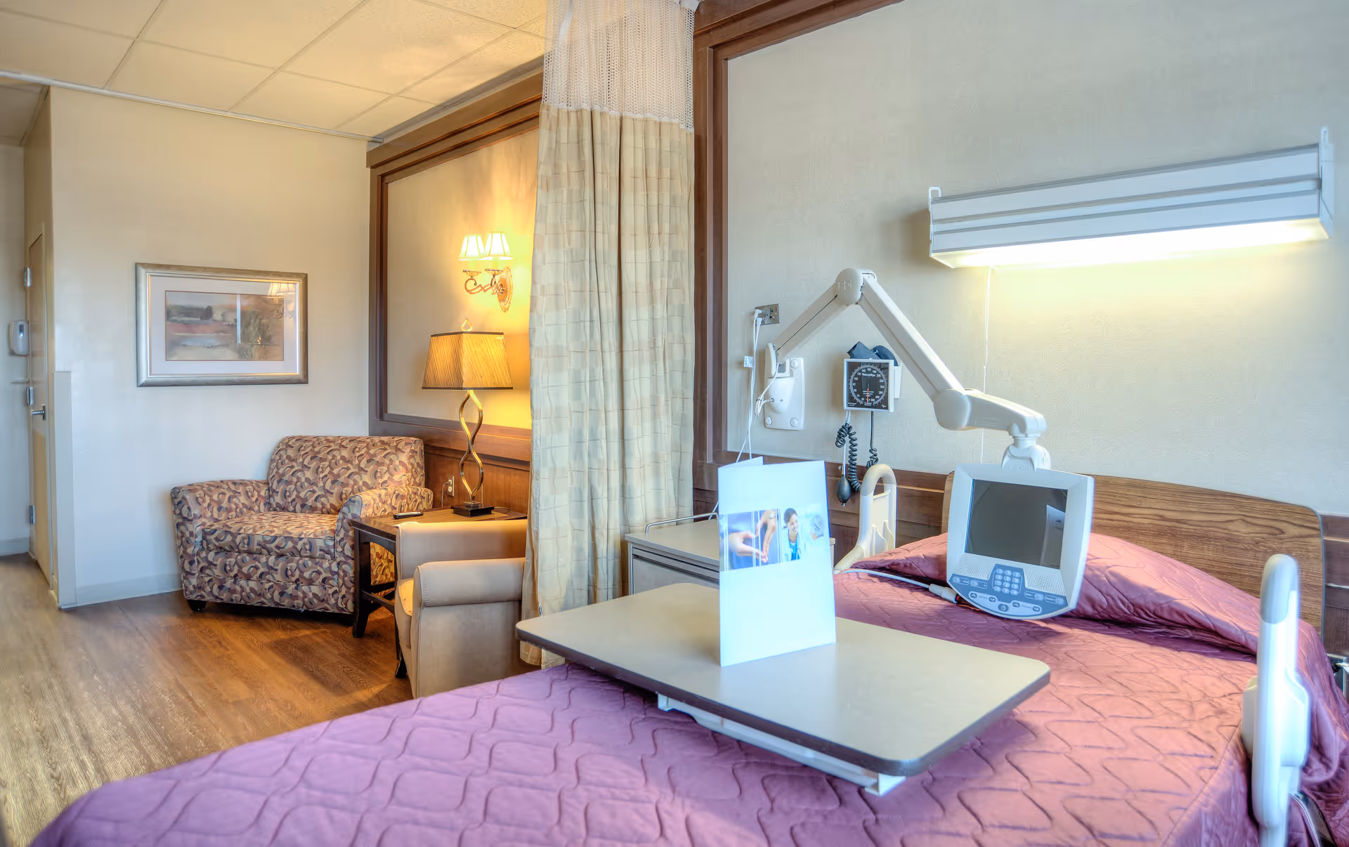 A hospital-style room with a bed covered in a purple quilt, a medical monitor attached to the headboard, and an adjustable overbed table. There is a privacy curtain partially drawn, a patterned armchair, a beige armchair, a side table with a lamp, and a framed picture on the wall. The floor is wooden, and the room has soft lighting.