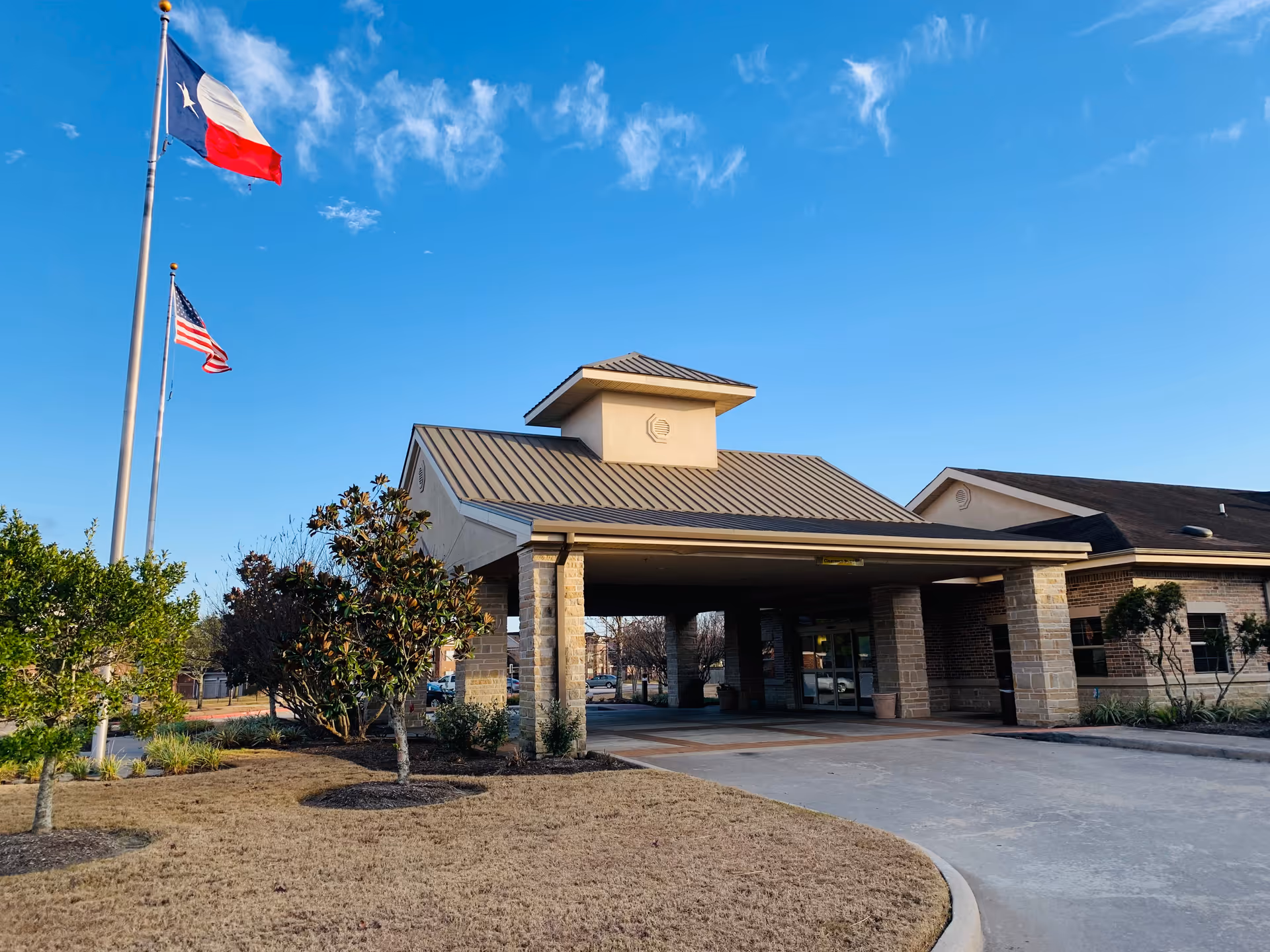 Exterior view of Falcon Point Post Acute building entrance with a covered driveway. Two flagpoles display the Texas state flag and the United States flag. The sky is clear and blue, and there are some trees and shrubs around the building.