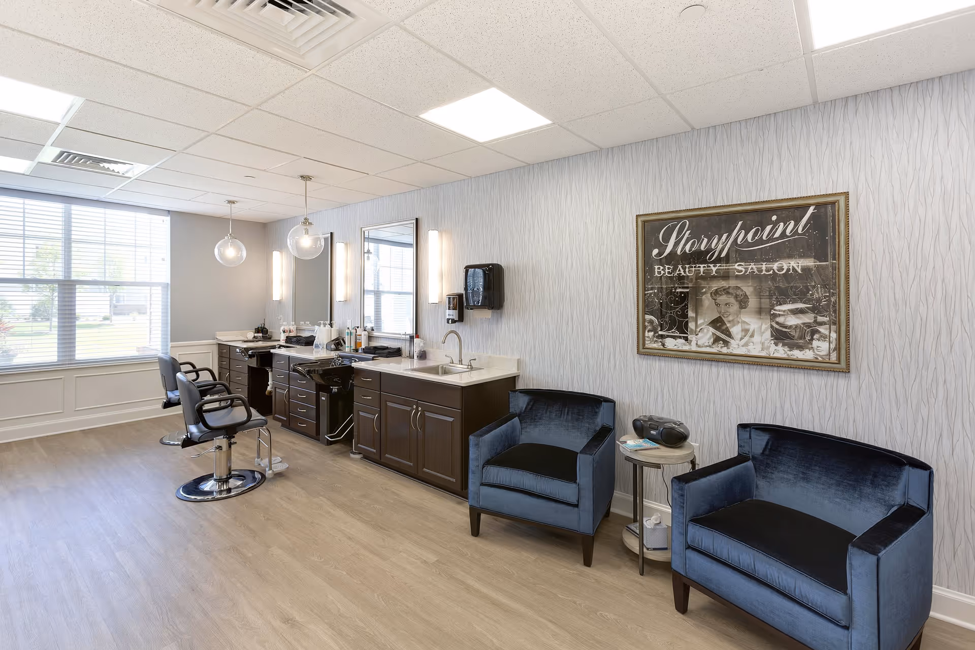 Interior of a beauty salon at StoryPoint Medina featuring two black salon chairs in front of large mirrors and a sink. The room has light wood flooring, a large window with blinds, two blue velvet armchairs, a small round table with a radio and magazines, and a framed vintage-style sign on the wall that reads 'Storypoint Beauty Salon'.