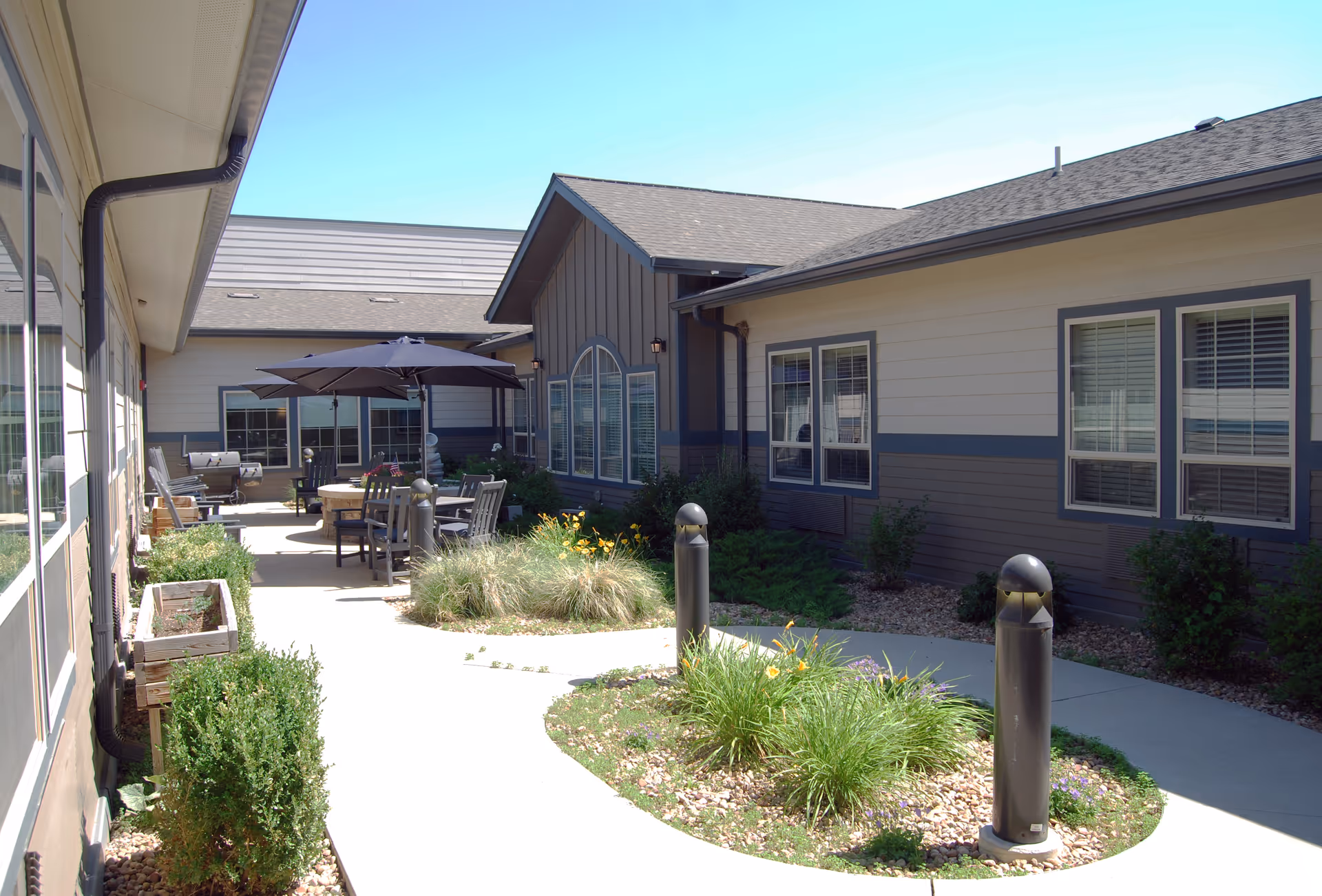 Outdoor courtyard area of a senior living facility with a curved concrete walkway, landscaped garden beds with plants and flowers, patio tables with umbrellas, and chairs. The building exterior features beige and gray siding with multiple windows under a clear blue sky.