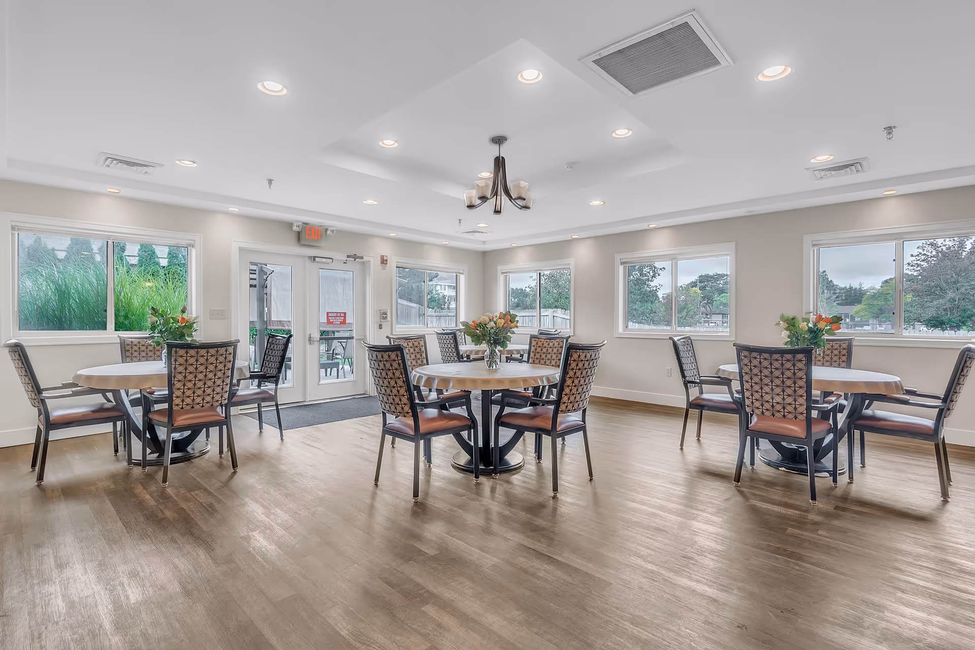 Bright communal dining room with round tables and chairs, large windows, and hardwood floors.