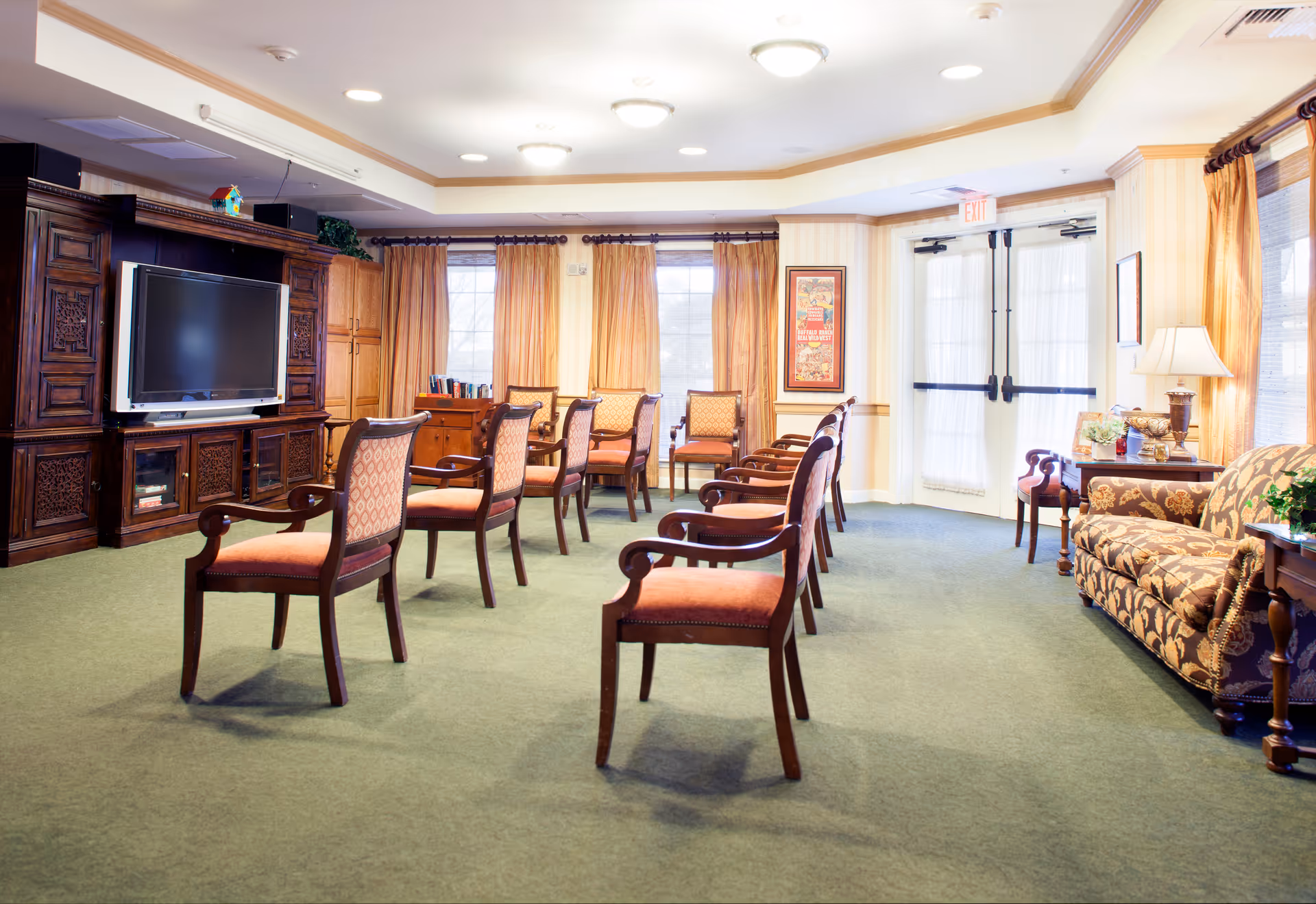 A bright communal living room with upholstered chairs arranged facing a large TV cabinet, a patterned sofa, windows with curtains, and double doors.