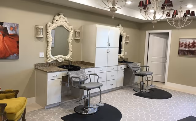 Interior view of a salon area in a senior living facility with two styling chairs in front of large ornate mirrors mounted on a beige wall. There are white cabinets and drawers between the mirrors, a chandelier overhead, and framed artwork on the walls.