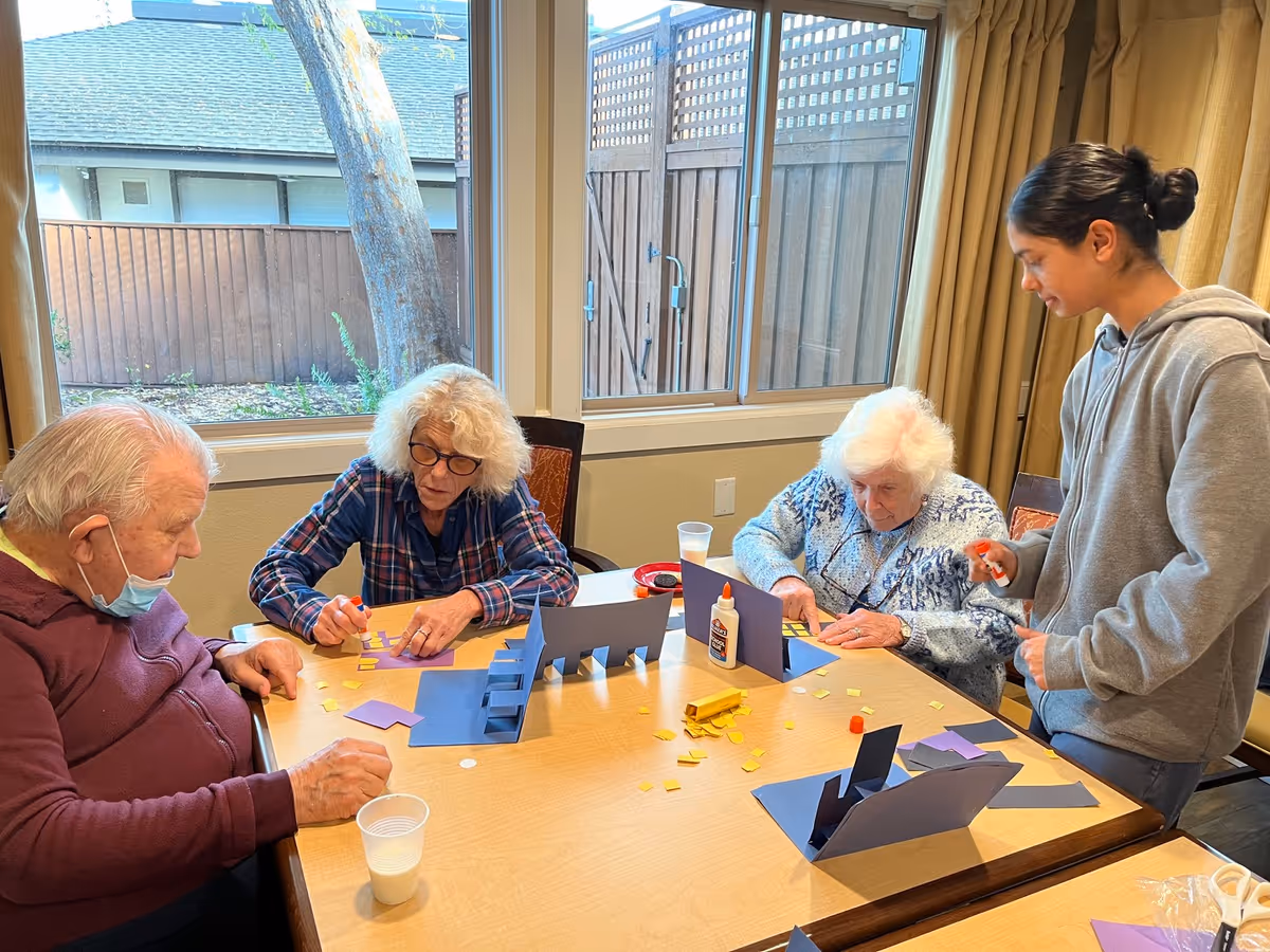 Three elderly individuals and a younger person engaged in a craft activity at a table inside a room with large windows showing an outdoor fence and tree. The table has glue, paper cutouts, and other craft supplies.