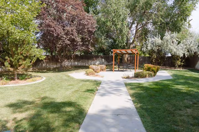 Concrete path leading through a grassy courtyard to a wooden pergola with seating, surrounded by trees and shrubs.