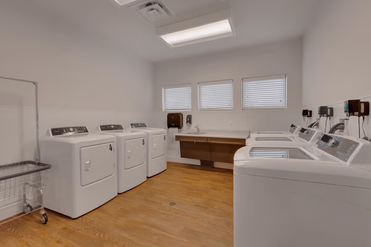 A laundry room with three white front-loading dryers numbered 4, 5, and 6 on the left side and three white top-loading washing machines on the right side. The room has wooden flooring, three small windows with blinds, a countertop with a sink, and a paper towel dispenser on the wall.