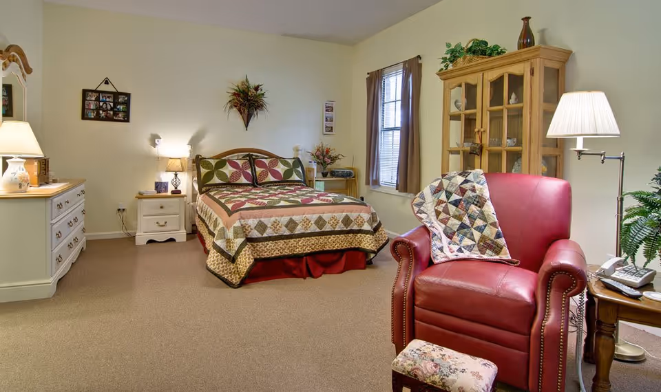 A cozy senior living bedroom with a bed covered in a colorful quilt, a red leather armchair with a patchwork quilt draped over it, a wooden cabinet with glass doors, a dresser with a lamp, and a window with brown curtains.