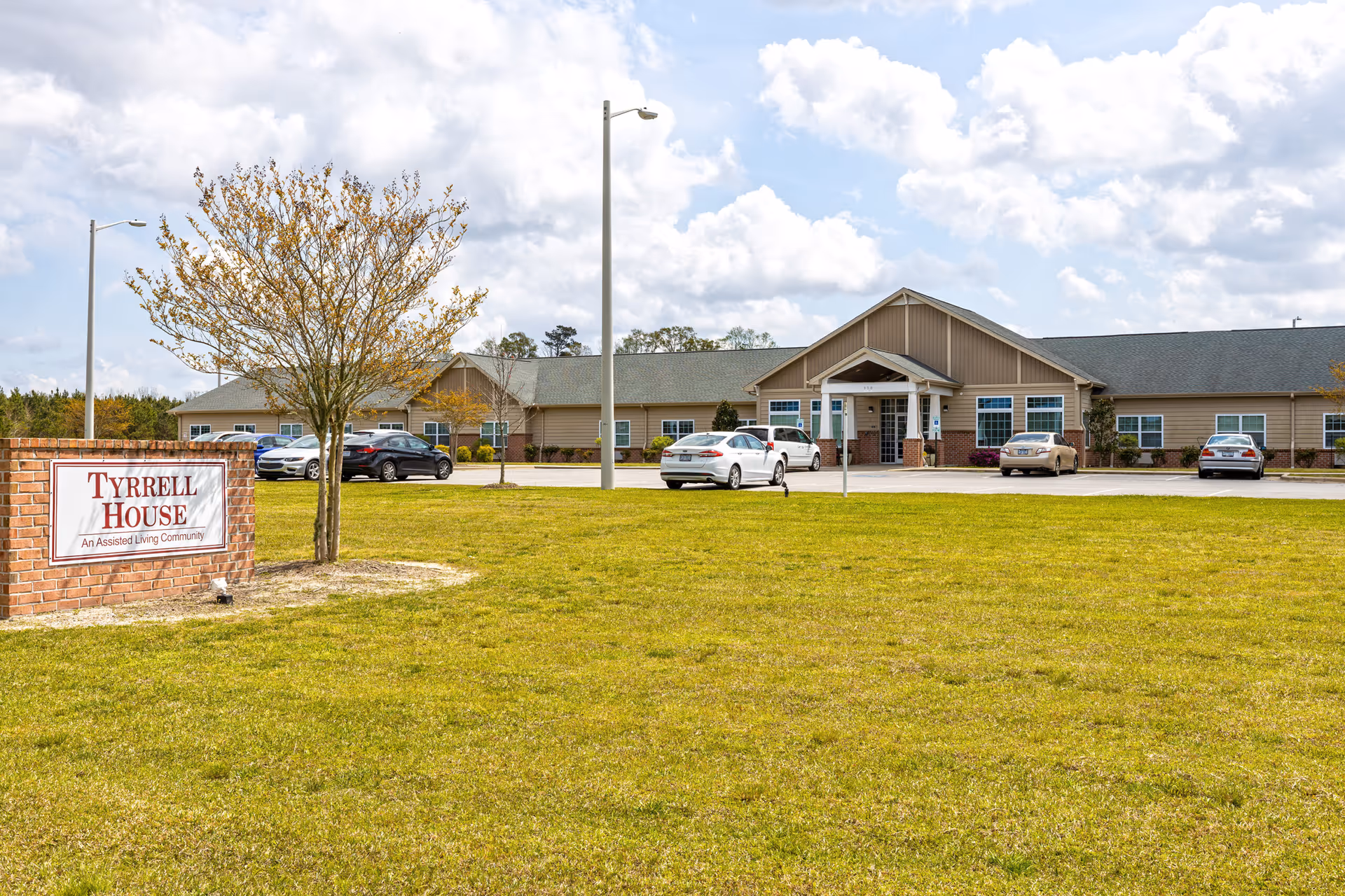 Exterior view of Tyrrell House, an assisted living community, showing a single-story building with a pitched roof, several windows, and a covered entrance. There are a few cars parked in front of the building, a grassy lawn, a tree, and a brick sign with the facility name.