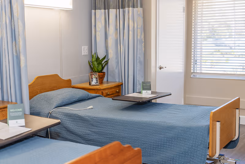 A room with two hospital-style beds covered with blue quilts, each with an overbed table holding a welcome card. There is a wooden nightstand between the beds with a potted plant and a framed photo. The room has light blue curtains and a window with blinds letting in natural light.
