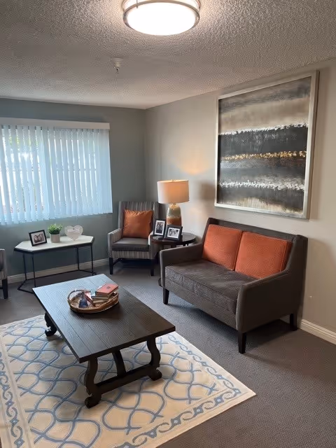 A cozy living room with a gray sofa featuring two orange cushions, a matching armchair with an orange pillow, a wooden coffee table on a patterned rug, a side table with a lamp and framed photos, a small table with a plant and decorative items near a window with vertical blinds, and a large abstract painting on the wall.
