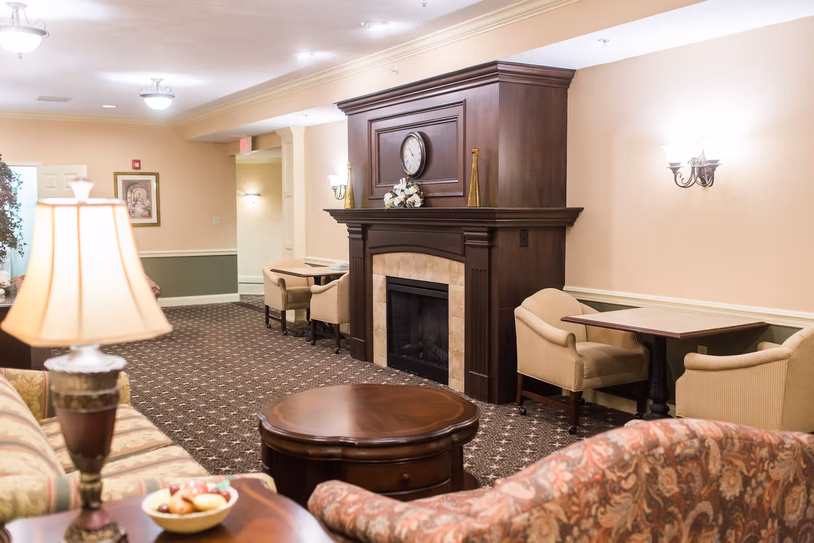 A cozy living room area in Briar Hill Health Care Residence featuring a dark wooden fireplace with a clock and decorative items on the mantle. The room has patterned carpet, beige walls with a green lower border, several upholstered chairs and sofas, a round wooden coffee table, and a table lamp. There are also small tables with chairs along the wall and soft lighting from wall sconces and ceiling fixtures.