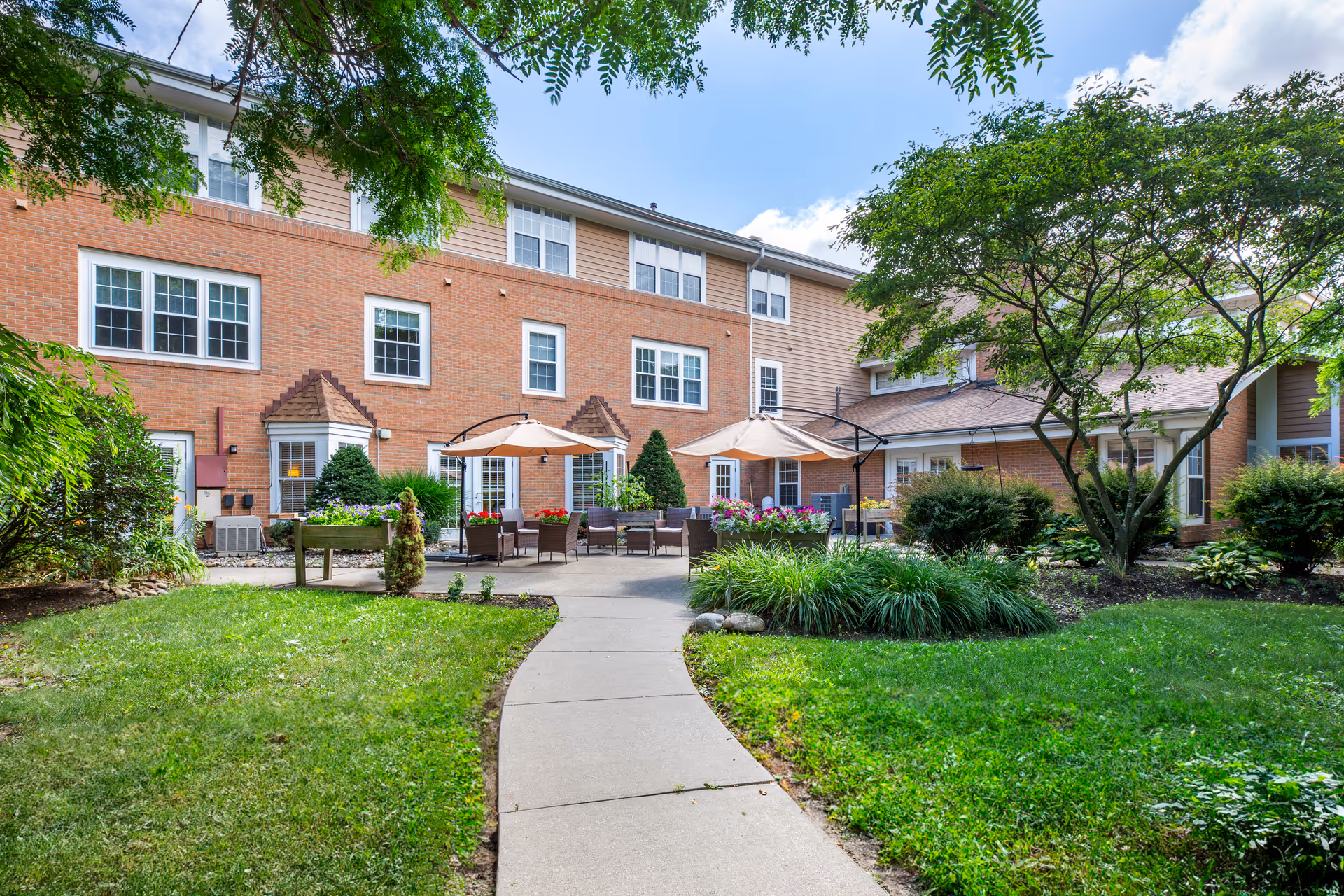 A landscaped courtyard with a paved walkway leading to patio seating, umbrellas, and a brick residential building.