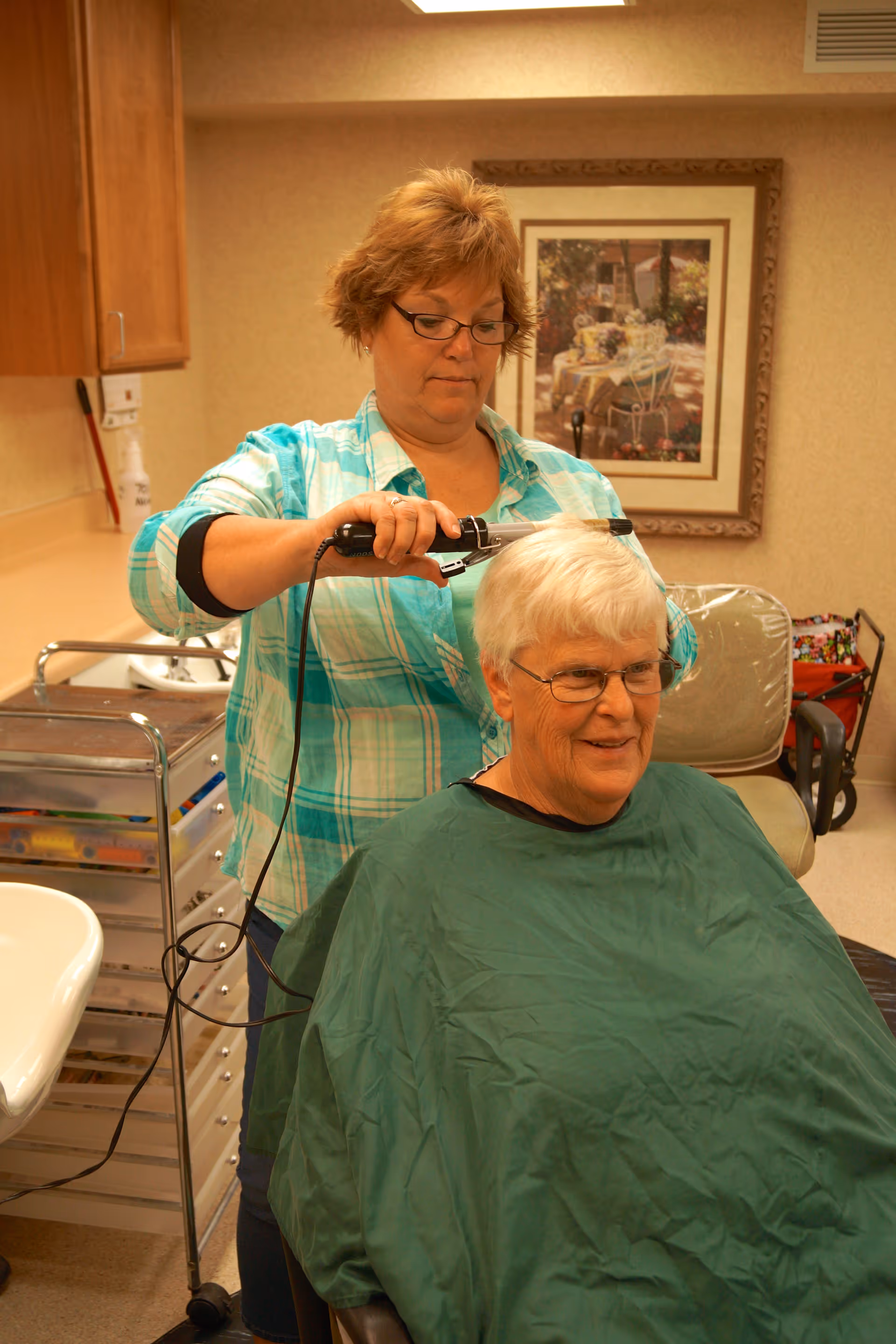 A woman wearing glasses and a plaid shirt is cutting the hair of an elderly woman with white hair who is seated and covered with a green cape in a salon-like room.