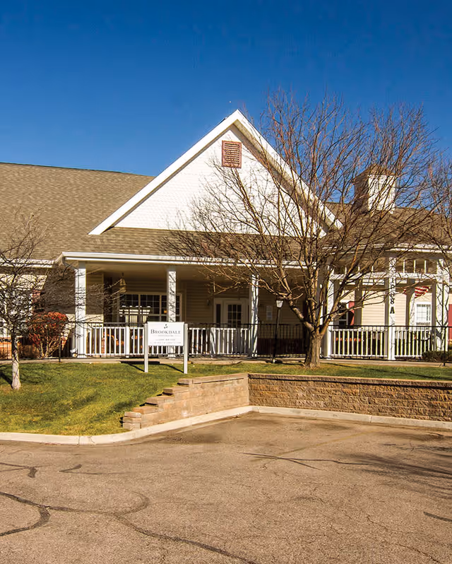 Exterior view of Brookdale Fort Collins Memory Care facility showing a single-story building with a porch, white railing, leafless trees, and a sign with the facility's name on a grassy area near a parking lot under a clear blue sky.