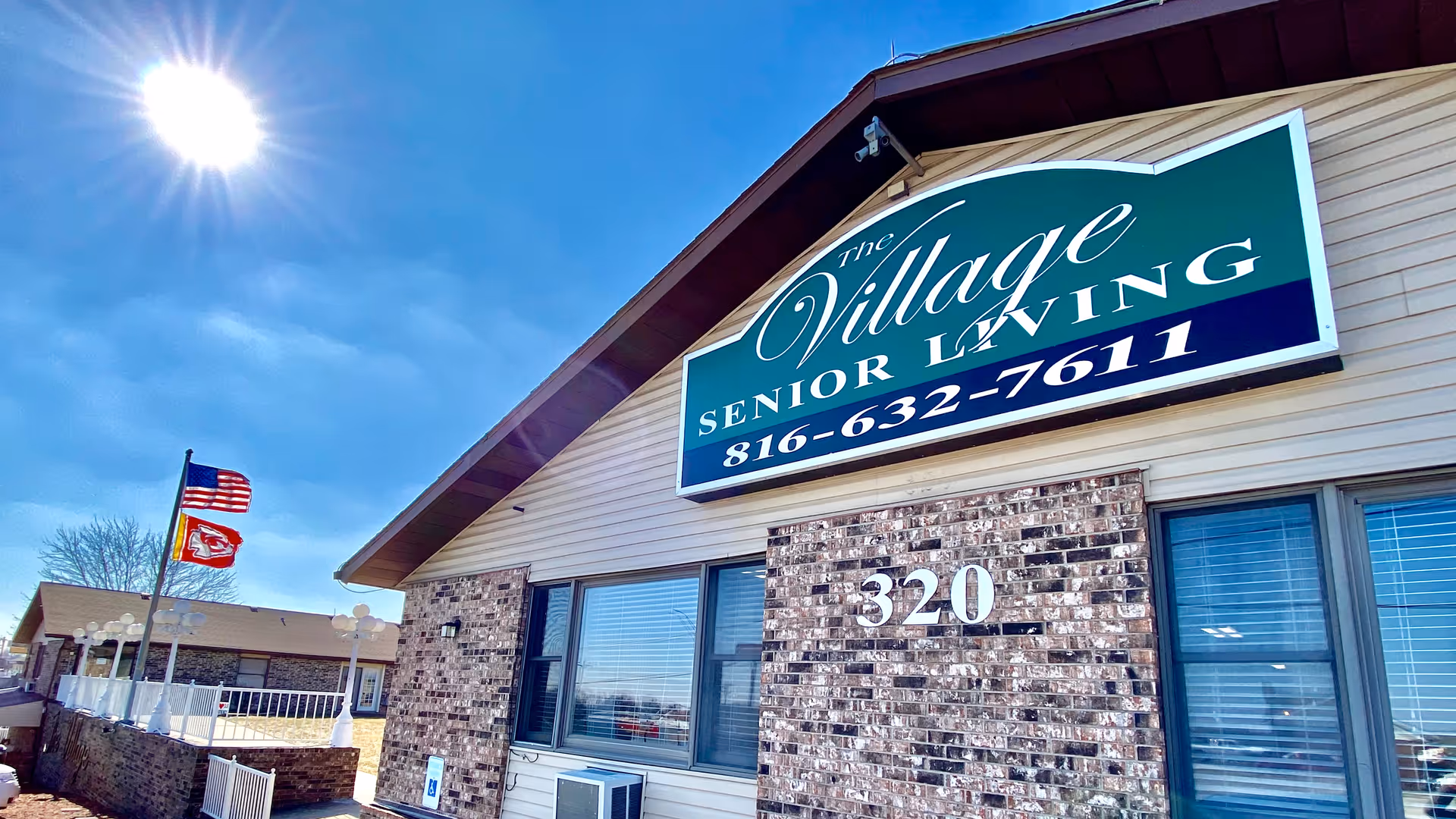 Exterior view of The Village Senior Living building under a bright blue sky with the sun shining. The building has a brick and siding facade with large windows and a sign displaying the facility name and phone number. Two flags, including an American flag, are flying on flagpoles near the building.