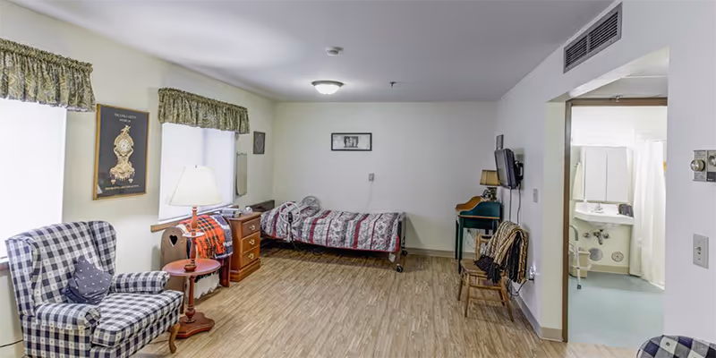 A senior resident room with a single bed, plaid armchair, dresser, and an open bathroom doorway.