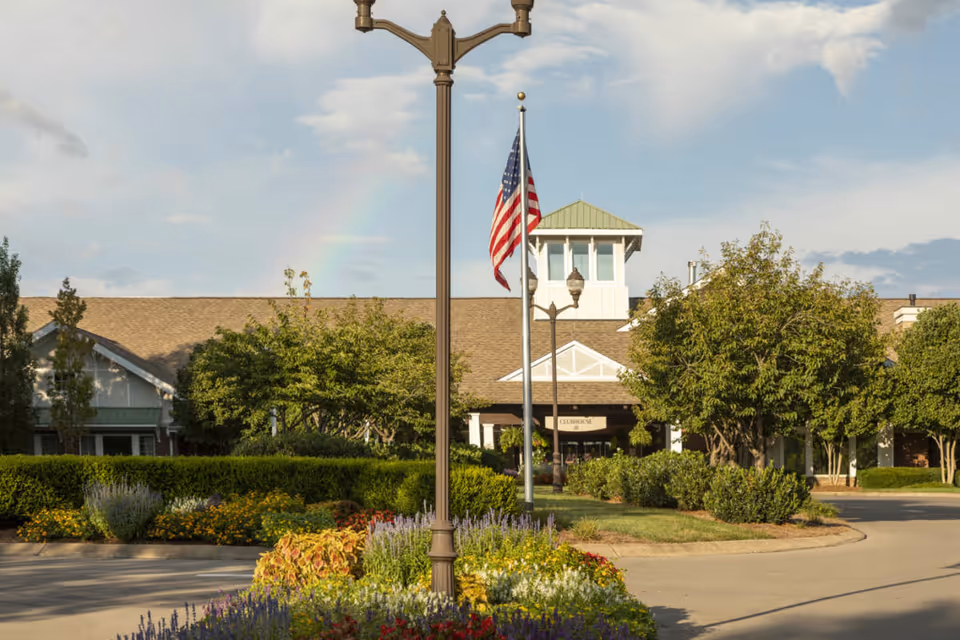 Exterior view of The Heritage at Brentwood facility with a landscaped garden featuring colorful flowers and bushes in the foreground, a tall street lamp, an American flag on a flagpole, and a building with a peaked roof and a small tower under a partly cloudy sky with a faint rainbow.