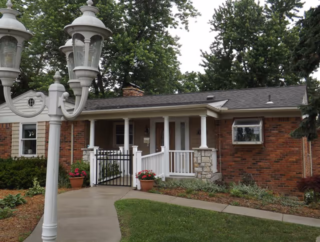 Single-story brick building front with a covered porch, white railings, a lamp post, walkway and potted flowers.